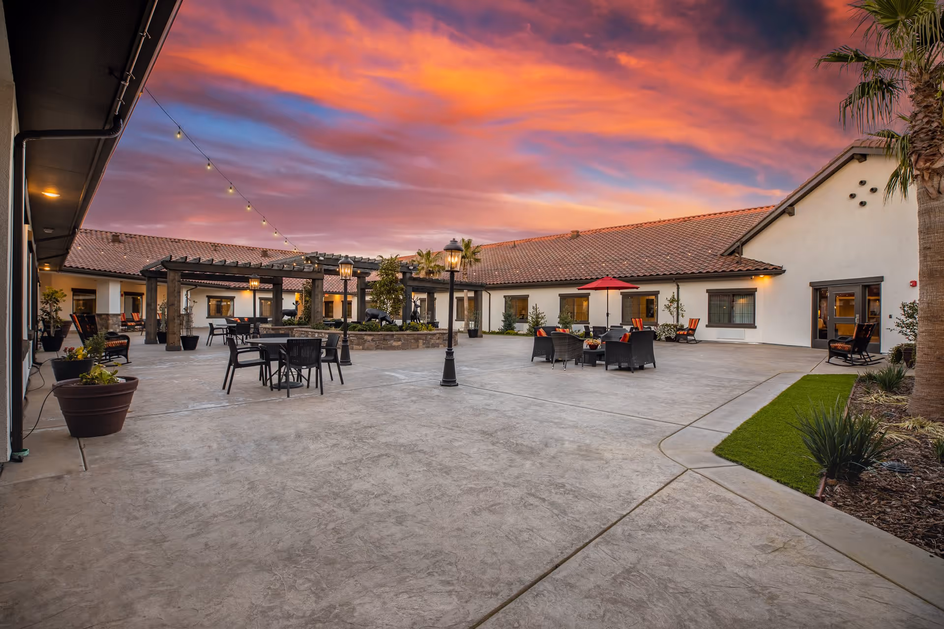 Outdoor courtyard area of The Gardens At Laguna Springs facility during sunset, featuring patio tables and chairs, a pergola, string lights, potted plants, and a vibrant colorful sky.