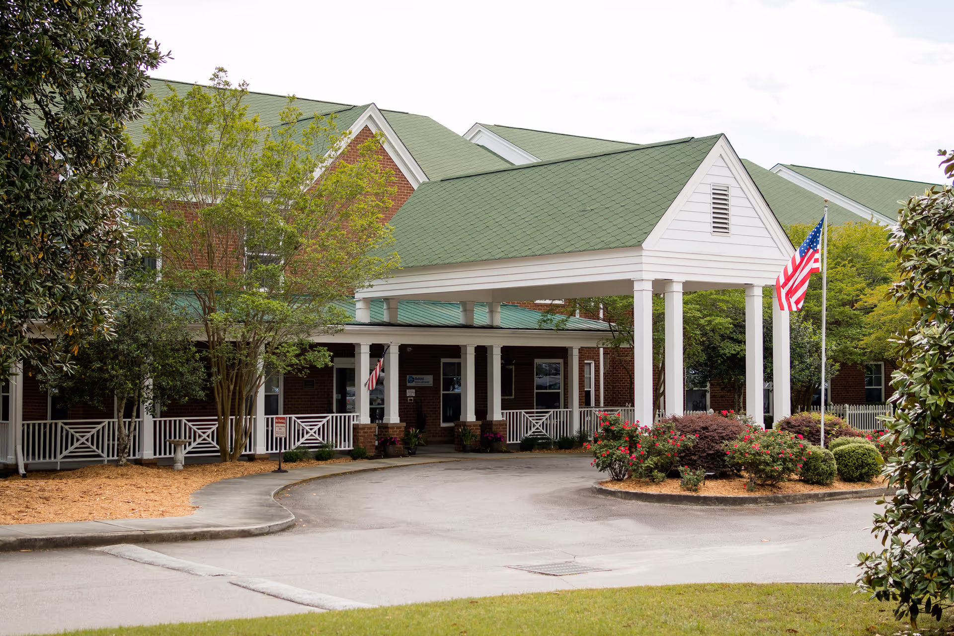 Entrance of a senior living facility with a green roof and white pillars supporting a covered driveway. There are trees and bushes with flowers around the driveway, and an American flag is displayed on a flagpole near the entrance.