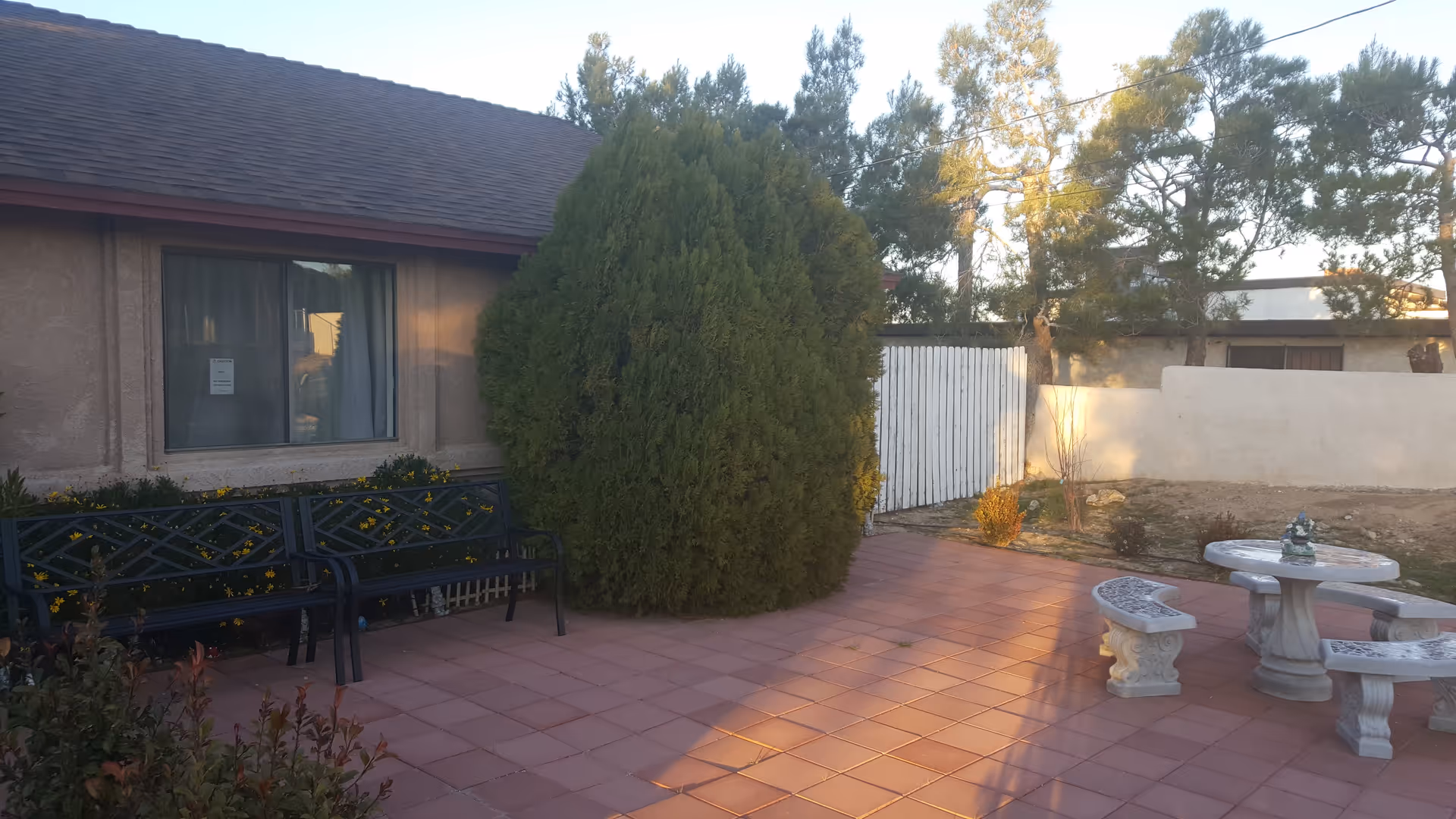 Outdoor patio area with tiled flooring, a large green bush, two black metal benches, a round stone table with decorative stone benches, and a beige building with a window in the background. Trees and a white wooden fence are also visible.