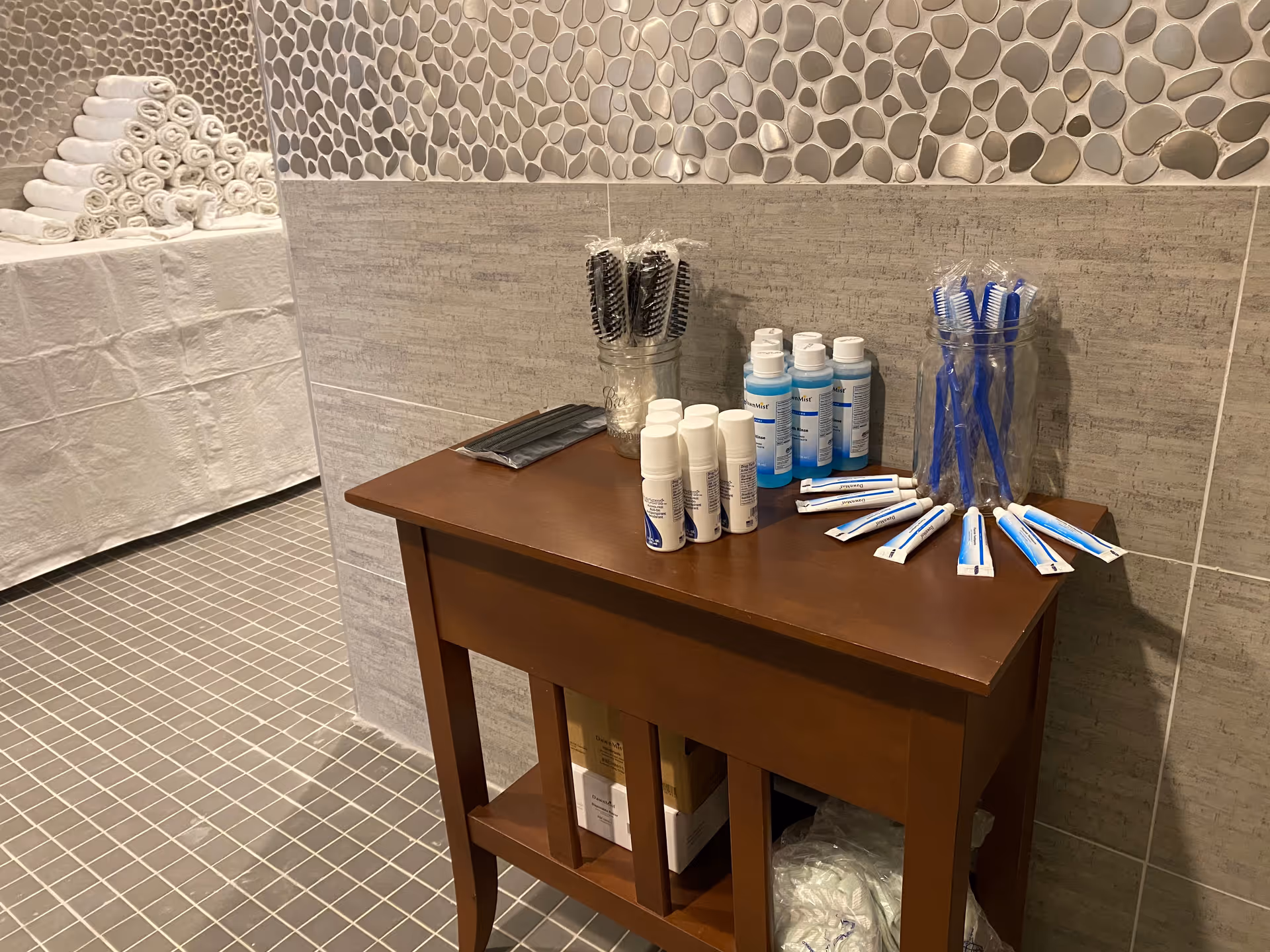 A small wooden table against a tiled wall holding personal hygiene items including toothbrushes, toothpaste tubes, mouthwash bottles, and hairbrushes. In the background, there is a stack of rolled white towels on a covered surface. The floor and walls are tiled, suggesting a bathroom or shower area.