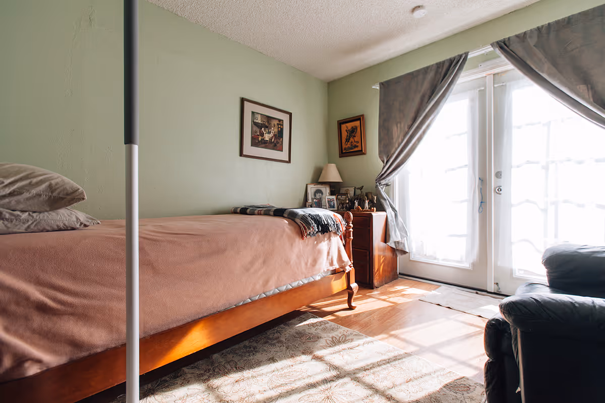 A cozy bedroom with a wooden bed covered in a brown blanket and pillows, a small wooden nightstand with a lamp and framed photos, two framed pictures hanging on a light green wall, and sunlight streaming through sheer curtains on glass doors. A dark armchair is partially visible on the right side.