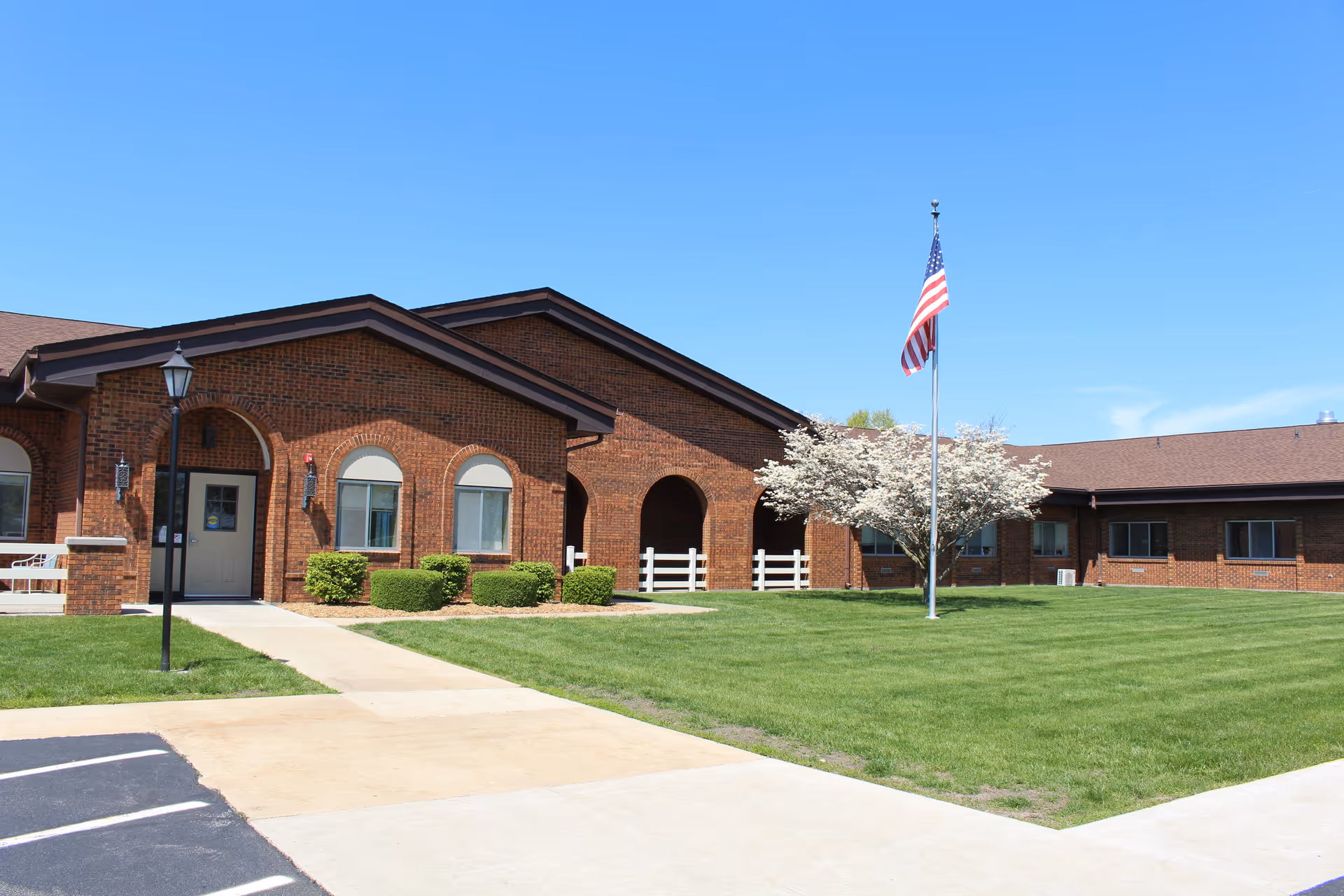 Front exterior of a brick senior living facility with an American flag, manicured lawn, and arched entrance.