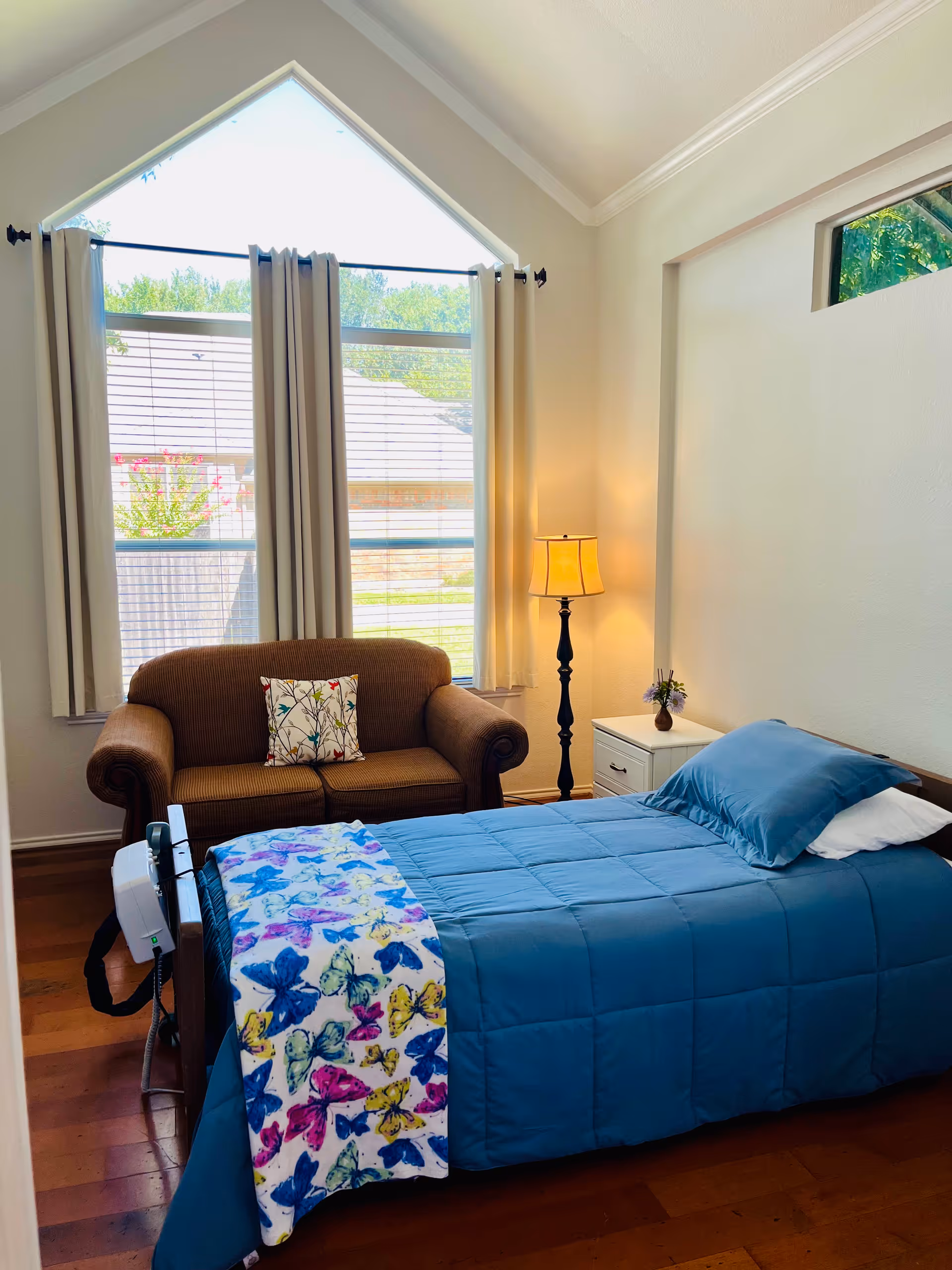 A cozy bedroom in an assisted living facility featuring a single bed with a blue comforter and a colorful butterfly blanket. Next to the bed is a white nightstand with a small vase of flowers and a tall floor lamp. A brown loveseat with a floral pillow is positioned in front of a large window with white curtains, allowing natural light to fill the room.