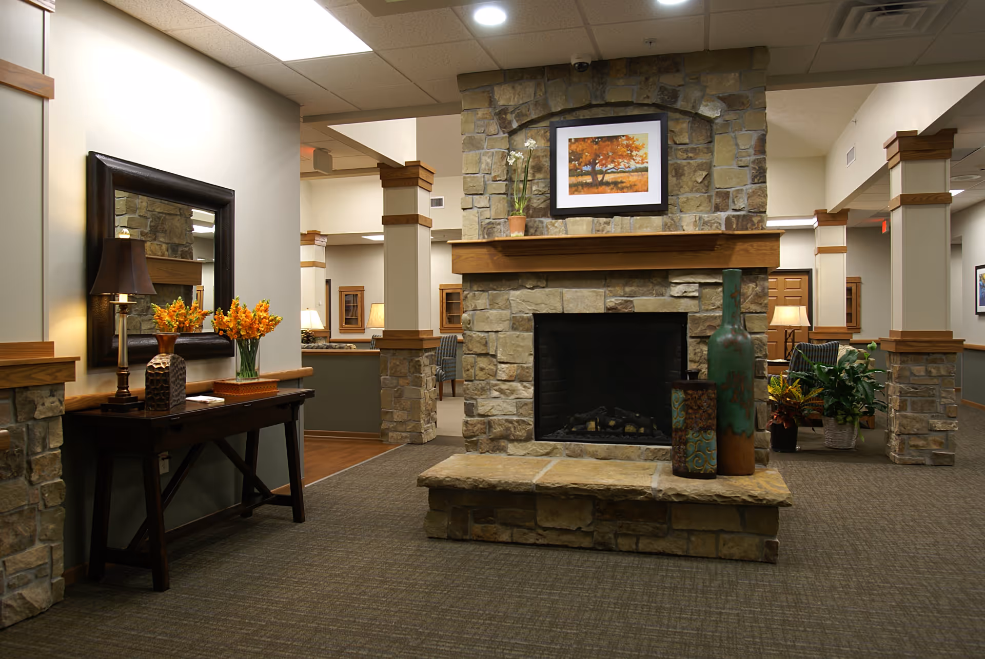 Interior view of a senior living facility lounge area featuring a large stone fireplace with a wooden mantle. On the mantle is a framed painting of an autumn tree and a potted plant. In front of the fireplace are decorative vases. To the left, there is a dark wooden console table with a lamp, a vase with orange flowers, and a large mirror above it. The room has carpeted floors, beige walls with wooden trim, and several columns with stone bases. Additional seating and lamps are visible in the background.
