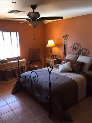 A cozy bedroom with a wrought iron bed frame, brown and beige bedding, a wooden nightstand with a lamp and picture frames, a wooden desk under a window with blinds, and a ceiling fan overhead.