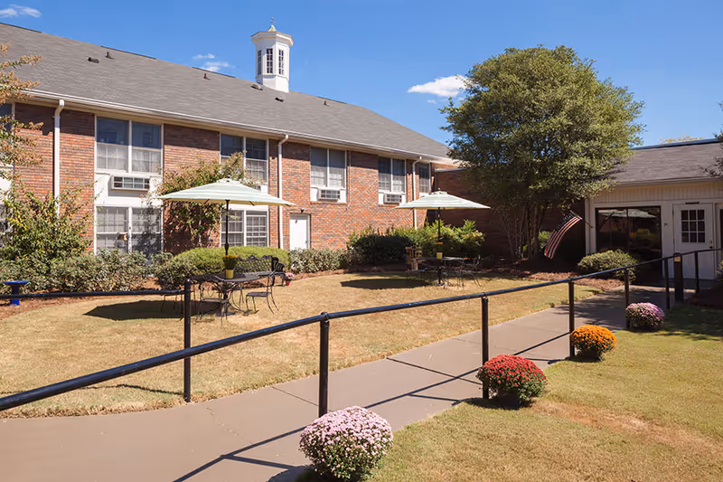 Sunny courtyard with brick building, patio tables and umbrellas, a paved walkway with handrails, and potted mums.