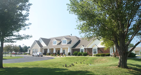 Front view of a single-story brick and siding senior living building with a driveway, parked car, manicured lawn, and trees.