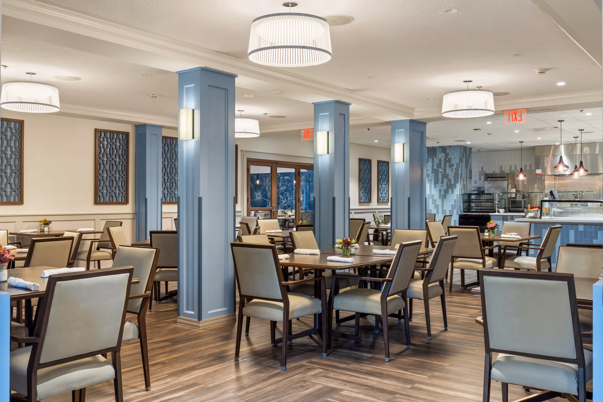 A spacious dining room with multiple tables and chairs arranged neatly. The room features light blue pillars, modern pendant lights, and decorative wall panels. The floor is wooden, and there is a serving counter with kitchen equipment in the background.