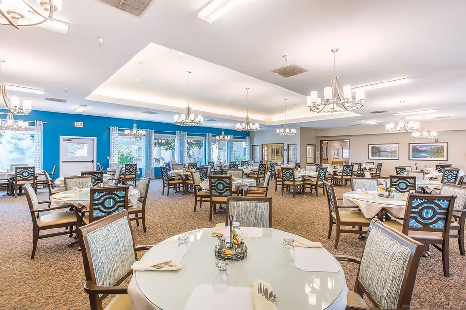 A spacious dining room with multiple round tables covered with white tablecloths, each set with napkins, glasses, and silverware. The chairs have patterned upholstery and wooden frames. The room features large windows with sheer curtains, blue accent walls, carpeted flooring, and several chandeliers hanging from the ceiling providing warm lighting.