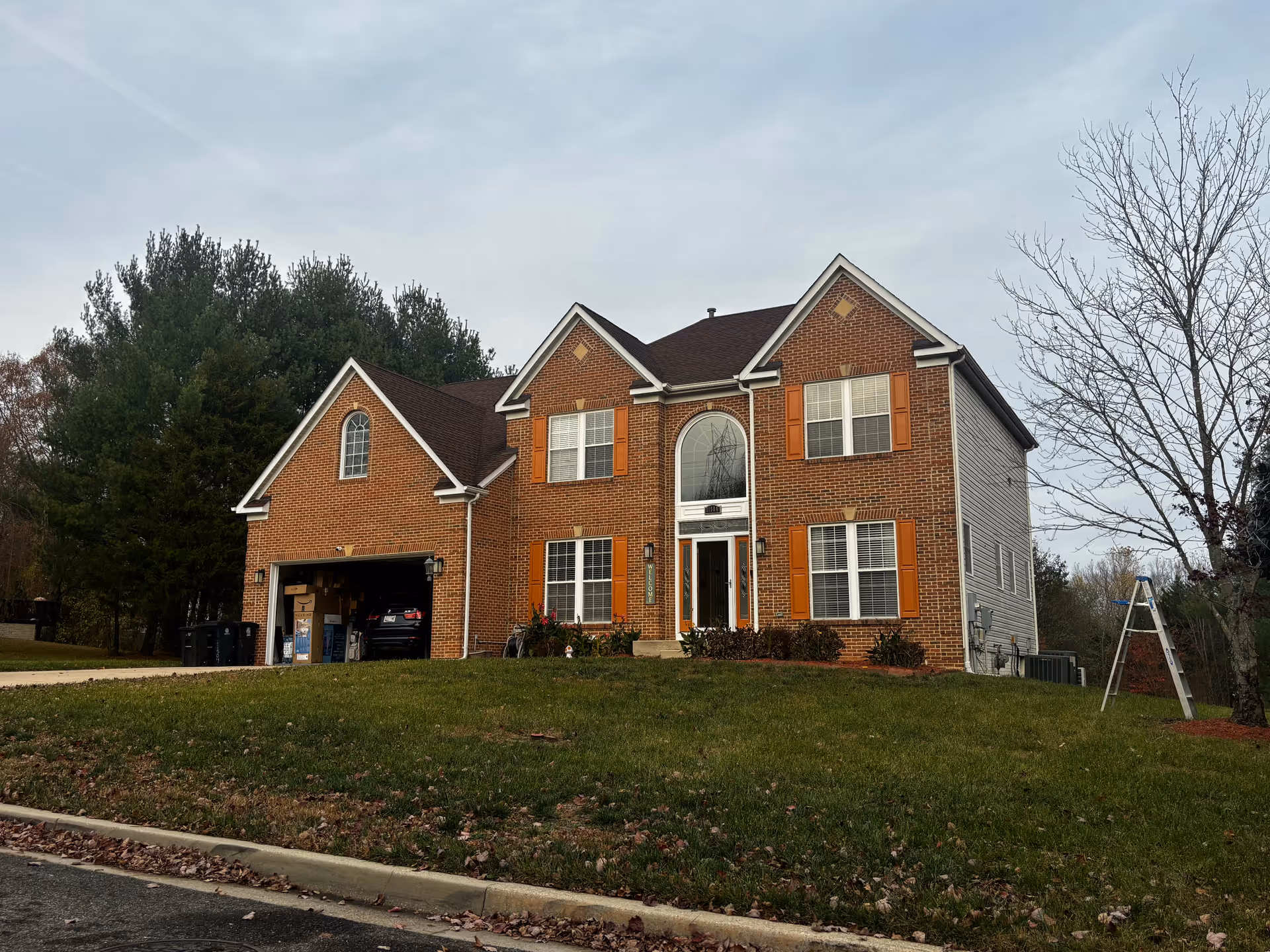 A two-story brick house with orange shutters and a large arched window above the front door. The garage door is open, showing a car and some boxes inside. The house is surrounded by a grassy lawn with some trees and a ladder on the right side. The sky is overcast.