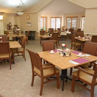 Well-lit communal dining room with several set wooden tables and cushioned chairs, each table featuring place settings and small floral centerpieces.