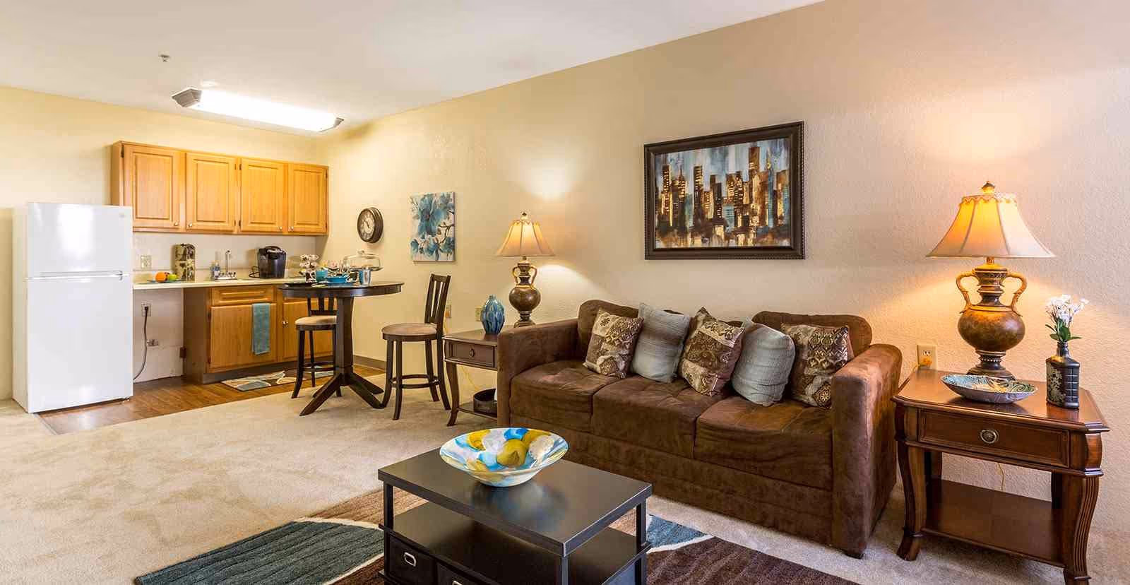 A cozy living area with a brown sofa adorned with multiple decorative pillows, a wooden side table with a lamp and a small plant, a coffee table with a decorative bowl, and a small dining area with a round table and two chairs. The background features a compact kitchen with wooden cabinets, a white refrigerator, and a countertop with a coffee maker and other items. The walls are decorated with a cityscape painting and a floral artwork.