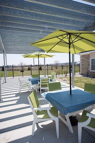 Outdoor patio area with tables covered by blue tablecloths and green umbrellas, surrounded by green and white chairs. The patio is shaded by a pergola and overlooks a fenced grassy area with a clear sky in the background.