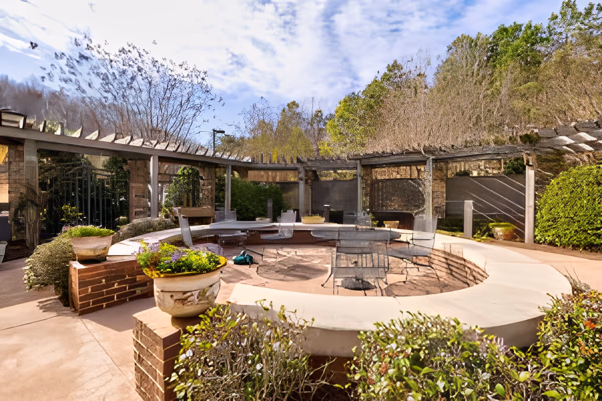 Outdoor circular seating area with a fire pit in the center, surrounded by plants and greenery. There is a pergola structure with wooden beams overhead and a partly cloudy sky in the background.