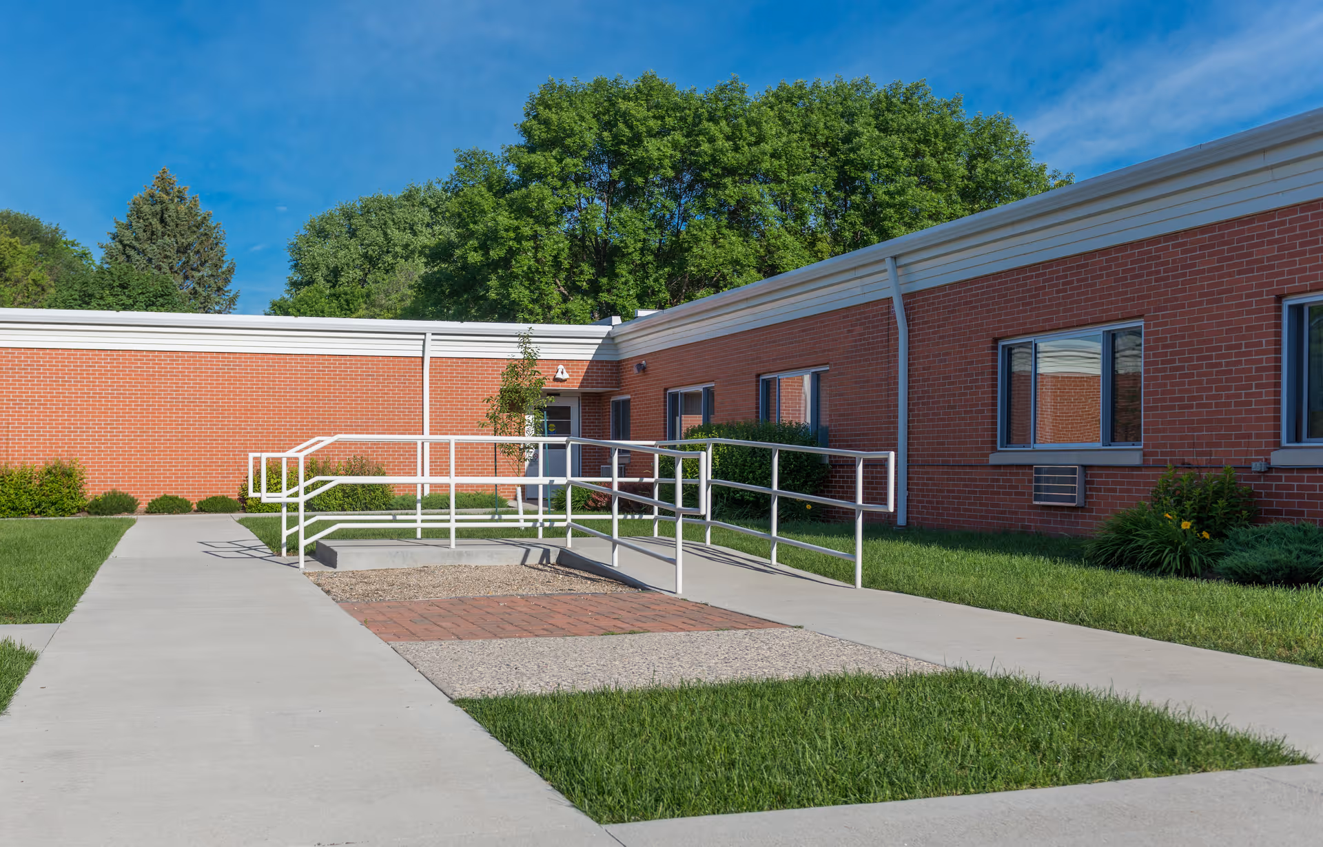 Exterior view of a single-story brick building with a wheelchair accessible ramp and handrails leading to a door. The building is surrounded by green grass, shrubs, and trees under a clear blue sky.