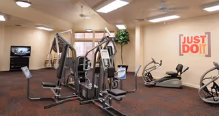 A fitness room with various exercise equipment including weight machines and stationary bikes. The room has beige walls, a carpeted floor, ceiling lights, and a large window letting in natural light. On one wall, there is a motivational sign that reads 'JUST DO IT'.