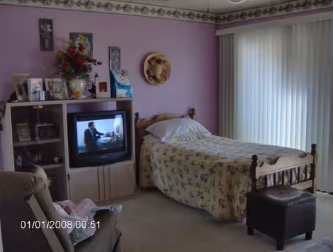 Cozy bedroom in a senior living facility with floral bedspread, TV, and large window blinds.