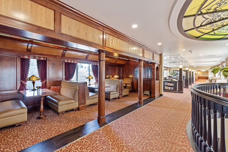 Wood-paneled dining alcoves with upholstered booths, stained-glass ceiling detail, patterned carpet and a long corridor in a senior living facility.