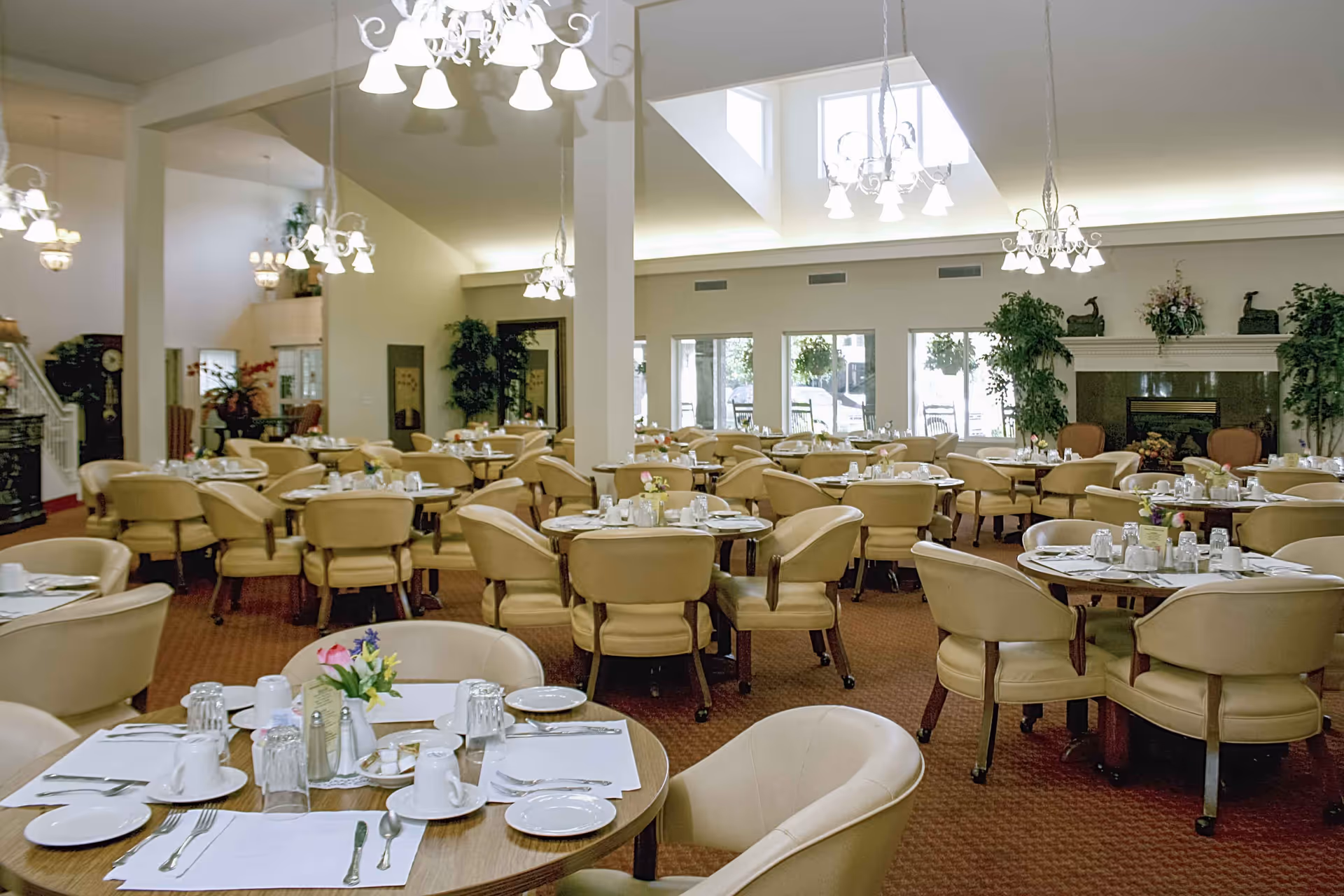 Spacious senior dining room with multiple round tables set for meals, beige chairs, chandeliers, plants, and a fireplace.