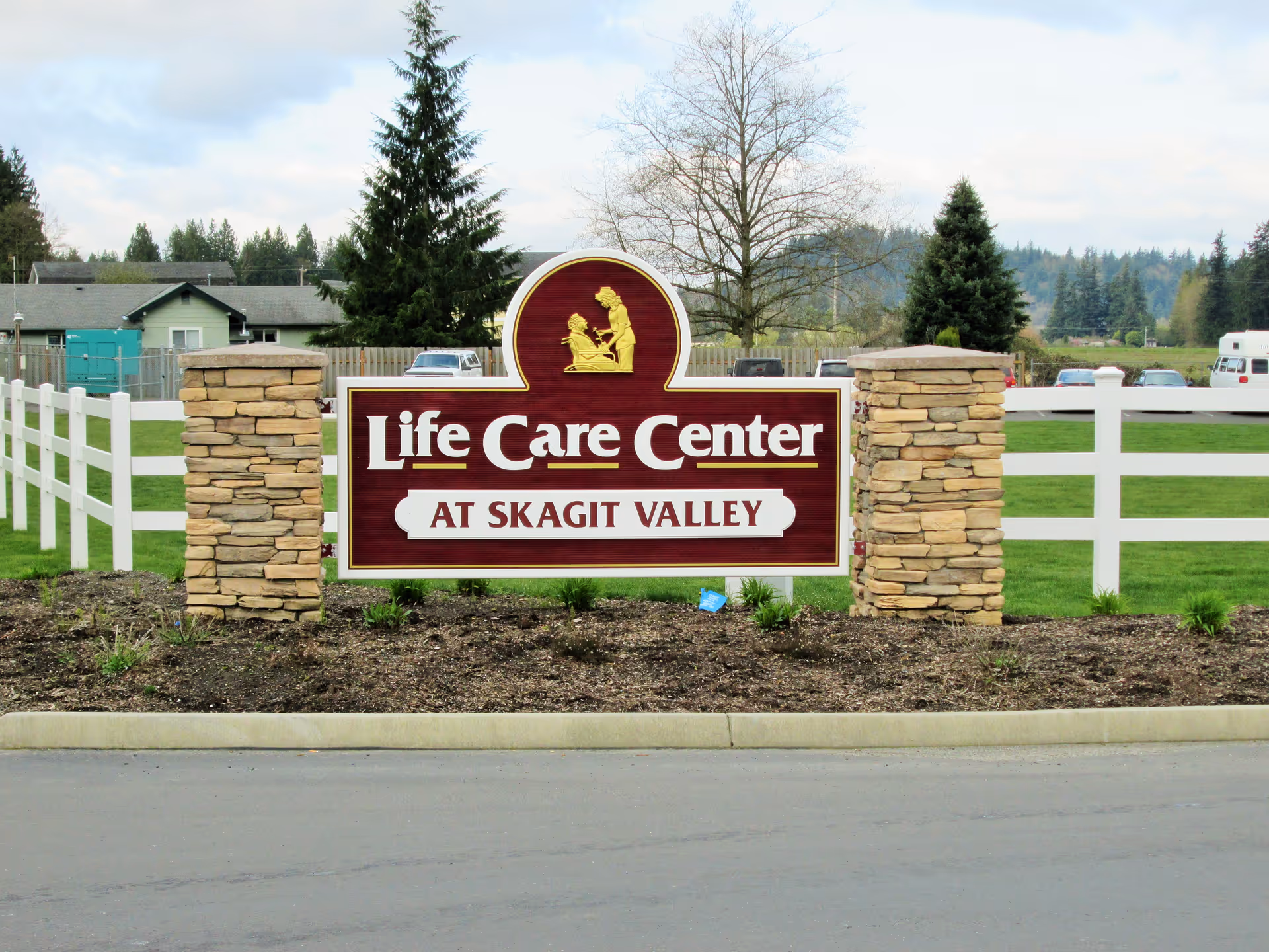 Outdoor view of a sign for Life Care Center at Skagit Valley, mounted between two stone pillars with a white fence and green grass in the background under a cloudy sky.