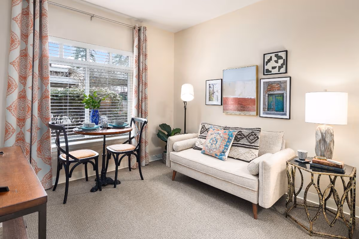 A cozy living room with a beige sofa adorned with patterned cushions, a small round table set for two with black chairs near a large window with patterned curtains, a floor lamp, a side table with a lamp and books, and framed artwork on the wall.