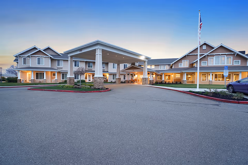 Front entrance of a two-story senior living building with a covered porte-cochere, flagpole, and parking area at dusk.