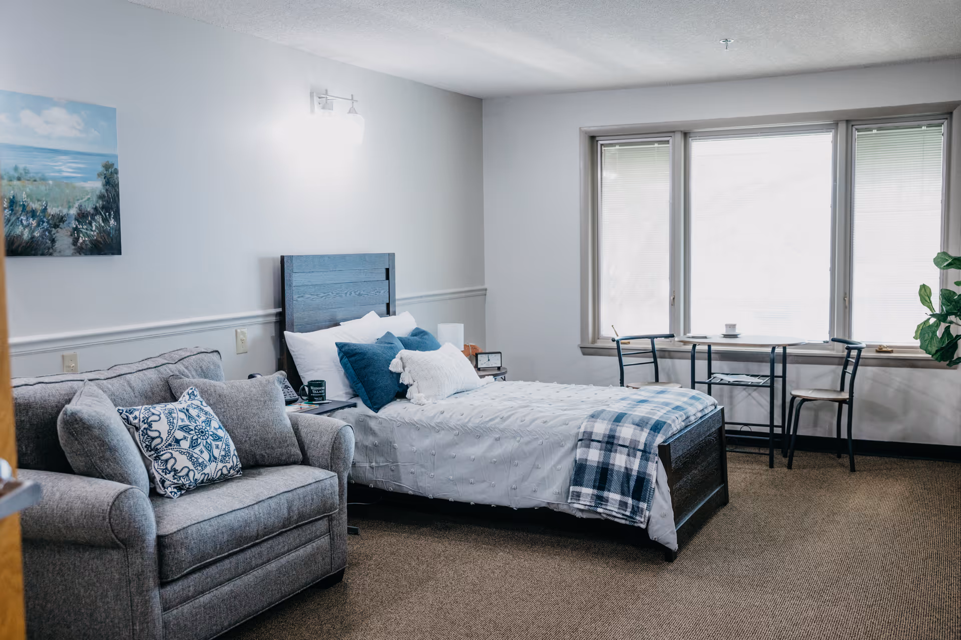 A cozy bedroom in an assisted living facility featuring a single bed with white bedding and blue pillows, a gray armchair with patterned cushions, a small table with two chairs by a large window, and a painting on the wall.