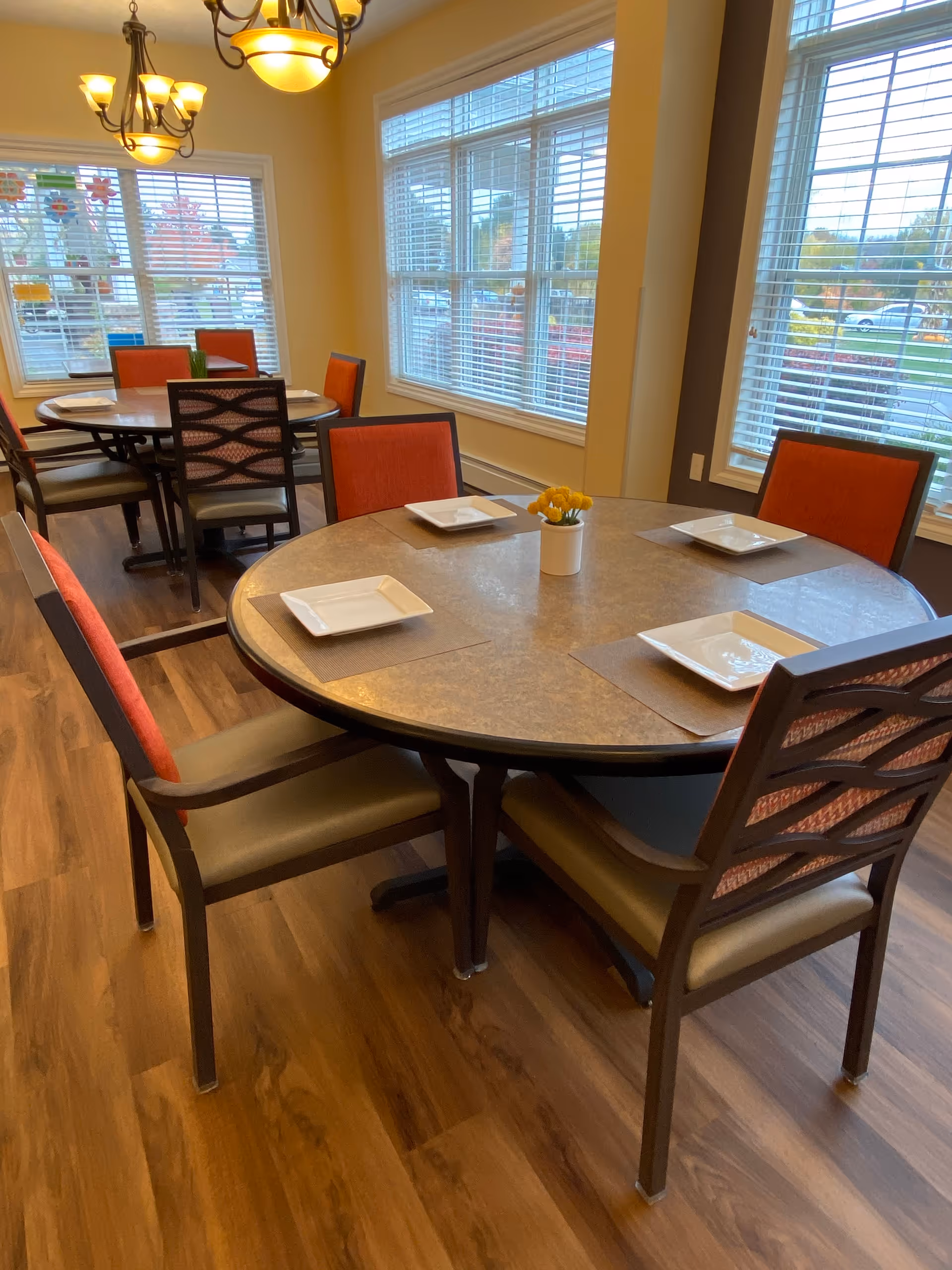 Bright dining room with round tables set with plates, orange-backed chairs, large windows with blinds, and wood flooring.