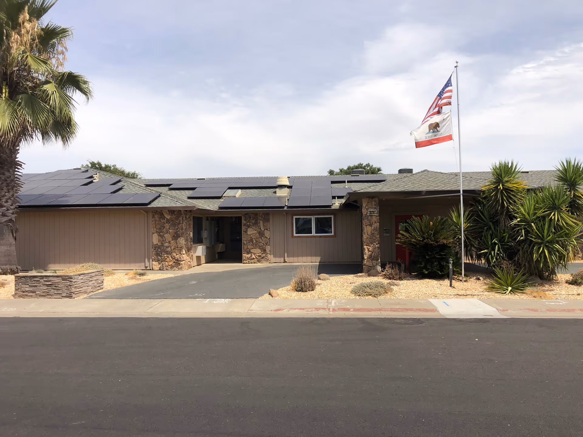 Single-story building with a stone and wood exterior, solar panels on the roof, an American flag and California state flag on a flagpole in front, surrounded by desert landscaping with palm trees and shrubs under a partly cloudy sky.
