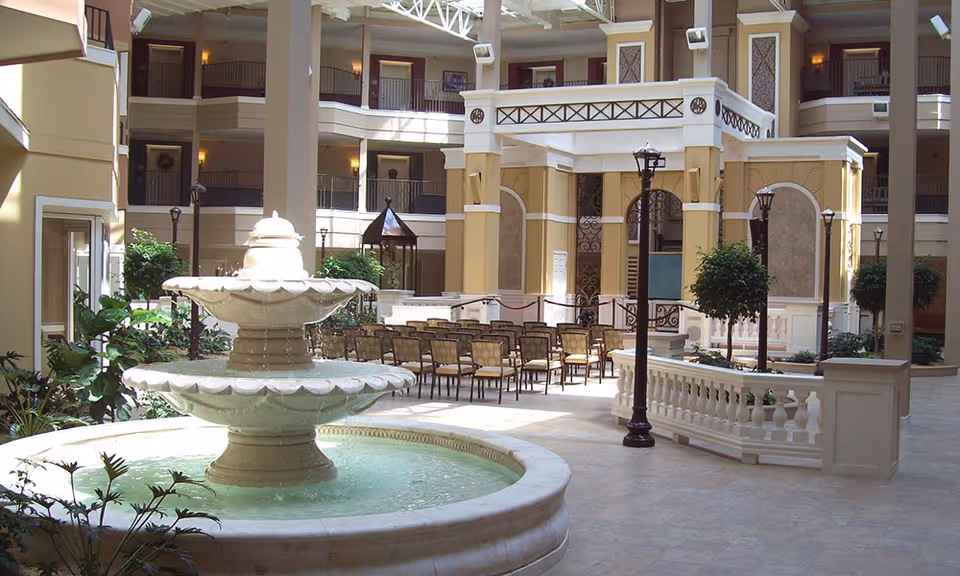Indoor atrium area with a large two-tiered water fountain in the foreground, surrounded by plants and decorative street lamps. In the background, there are rows of chairs arranged facing a small stage or podium area, with balconies and railings on multiple floors of the building visible above.