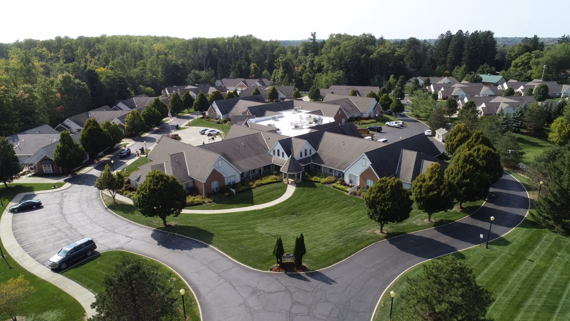 Aerial view of Wedgewood Estates senior living facility showing multiple single-story buildings with pitched roofs arranged around a central green lawn and curved driveway. The area is surrounded by trees and greenery, with several parked cars visible along the driveway.