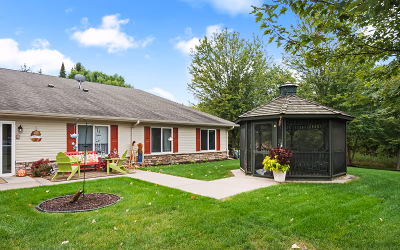 Outdoor view of Encore Assisted Living and Memory Care at Hugo showing a single-story building with beige siding and red shutters, a green lawn with a concrete walkway, a small garden bed with mulch, two green Adirondack chairs with a small table, and a dark green screened gazebo surrounded by trees under a partly cloudy blue sky.