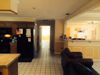Interior view of a senior living facility showing a kitchen area with dark wooden cabinets and a black refrigerator on the left, a hallway in the center leading to another room with natural light, and a reception or common area with a counter and bulletin boards on the right. There is a brown armchair in the foreground on the right side.
