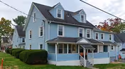 A light blue two-story residential building with a covered front porch and multiple dormer windows on the roof. The house is surrounded by a neatly trimmed hedge and a grassy lawn. There are other houses visible in the background along the street.