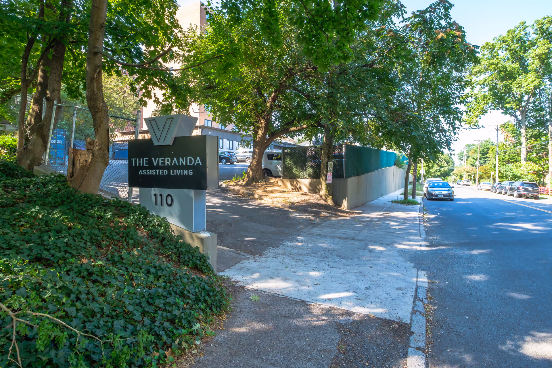 Sign reading 'The Veranda Assisted Living 110' at the facility entrance beside a tree-lined sidewalk and street.