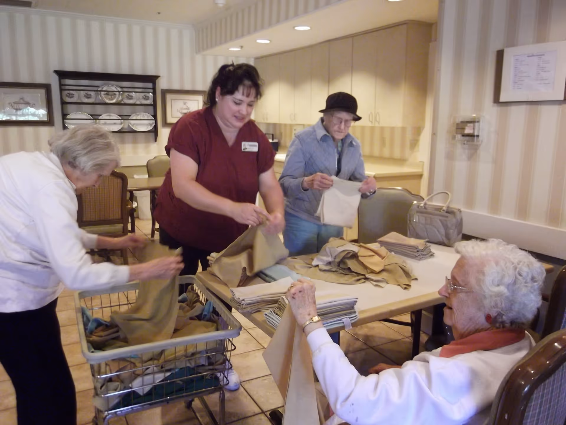 Four elderly women and a caregiver folding and sorting laundry in a room with beige striped wallpaper, a table, chairs, and cabinets in the background.