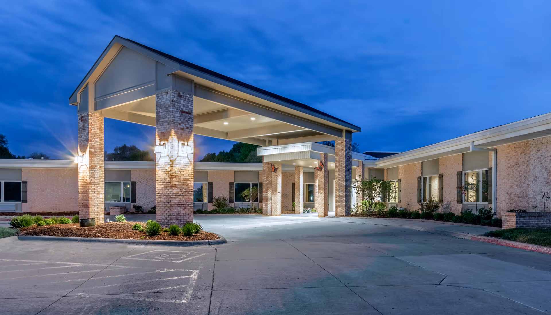 Exterior view of Brookestone of Papillion senior living facility at dusk, showing a covered entrance with brick pillars and well-lit facade surrounded by landscaped greenery.