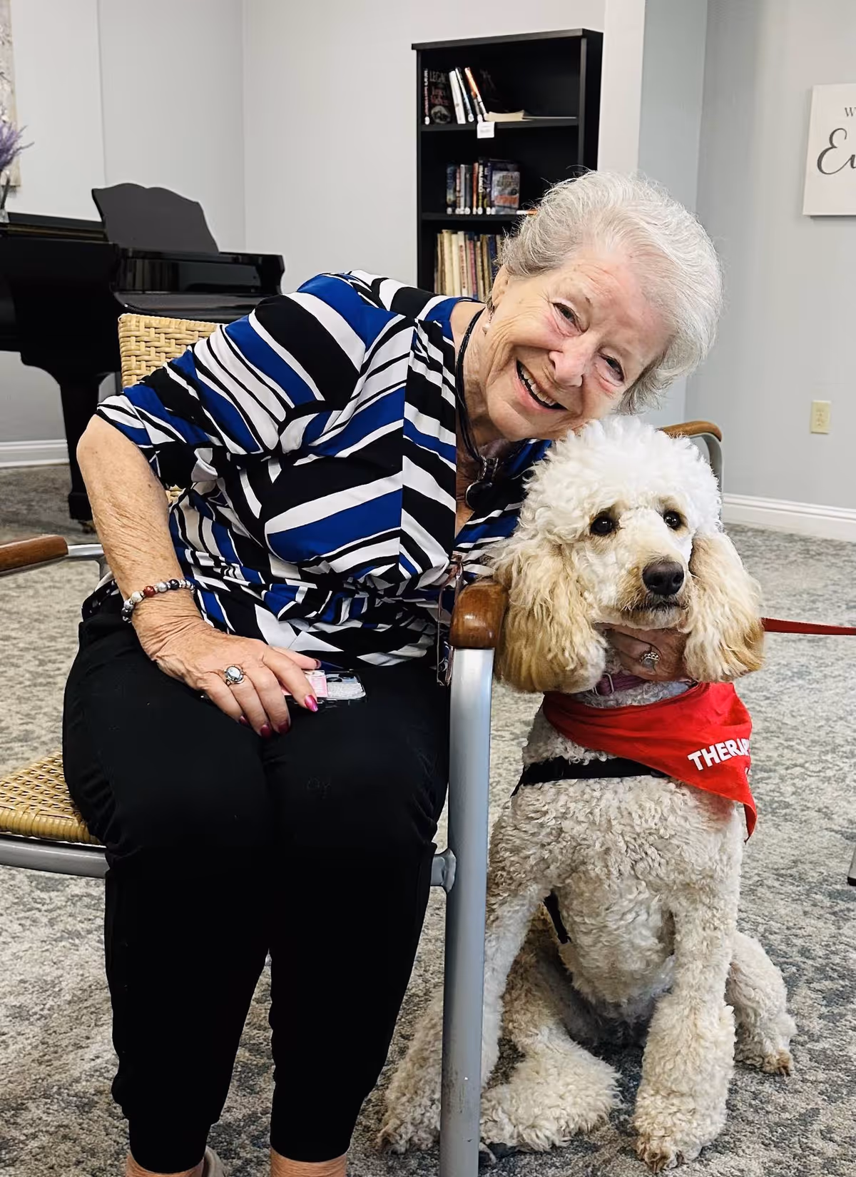 An elderly woman smiling while sitting next to a therapy dog in a cozy interior setting.