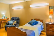 A cozy single bedroom with a bed, blue recliner, nightstand, desk, and warm wall lighting.