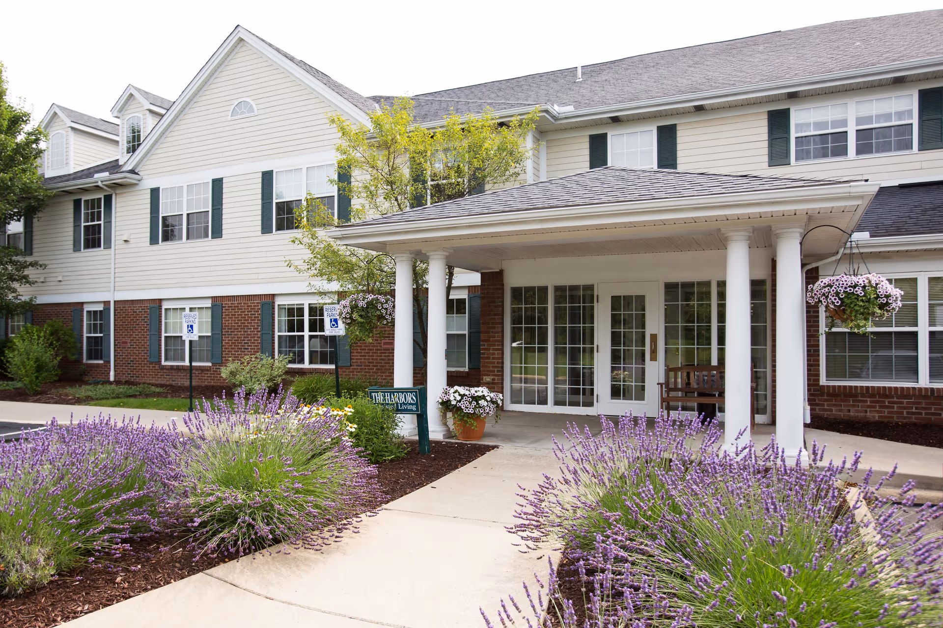 Exterior view of a senior living facility entrance with a covered porch supported by white columns. The building has beige siding with green shutters and brick accents. There are purple flowering plants and greenery along the walkway leading to the entrance. Two handicap parking signs are visible near the building.