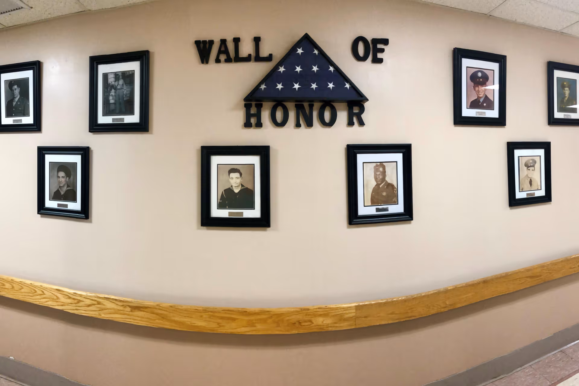 Indoor hallway 'Wall of Honor' display with a folded American flag, framed veteran portraits, and a wooden handrail.