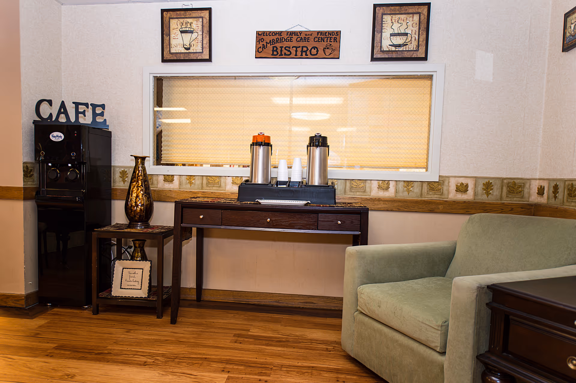 A small indoor cafe/bistro area with coffee dispensers on a table, a 'CAFE' sign, decorative vases, and a green armchair.