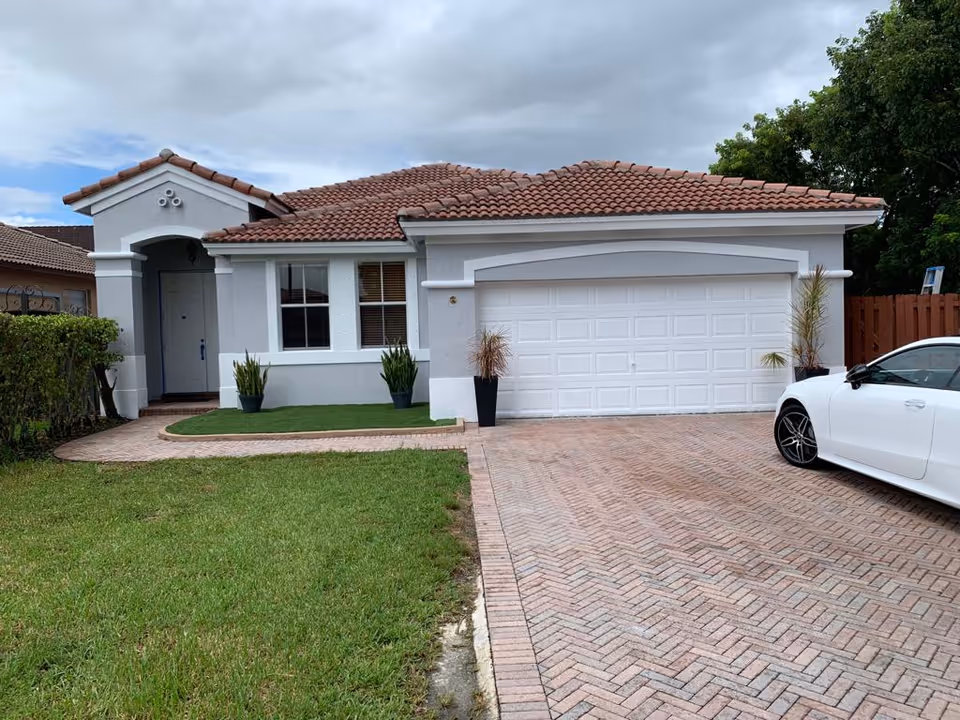 Single-story stucco house with a tiled roof, two-car garage, brick driveway, lawn, and a white car parked at the right.