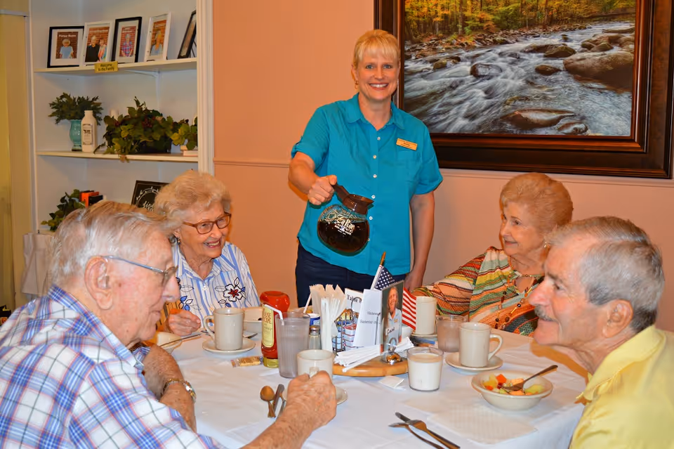 A staff member pours coffee for a group of elderly residents seated around a table in a dining room.