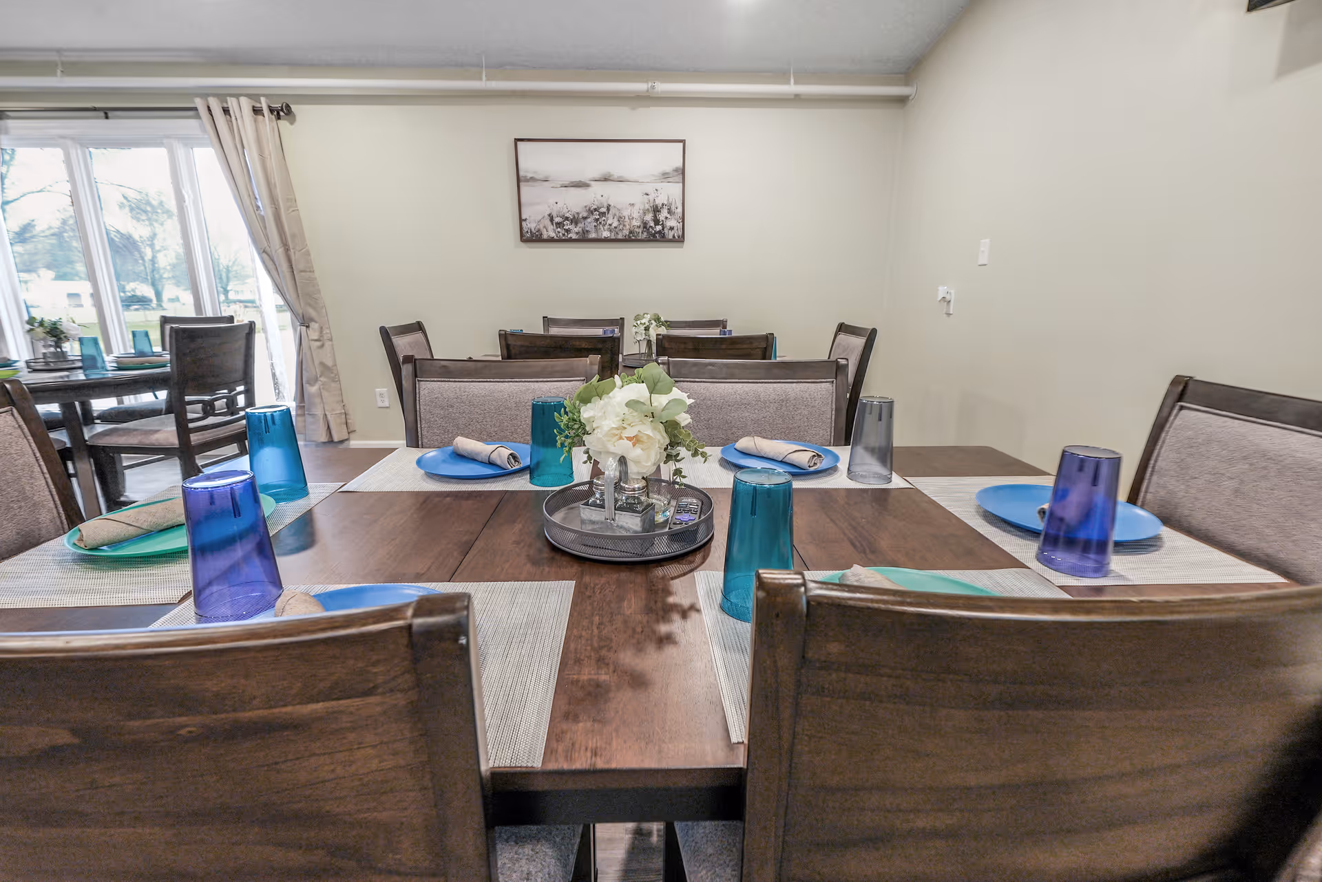 A dining room with a wooden table set for six people, featuring blue and green plates, rolled napkins, and blue and gray drinking glasses. A small floral centerpiece is placed in the middle of the table. The room has beige walls, a framed picture on the wall, and a large window with beige curtains letting in natural light.