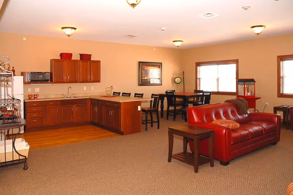 A senior living facility common area featuring a small kitchen with wooden cabinets, a microwave, and a sink. Adjacent to the kitchen is a dining area with tables and chairs. In the foreground, there is a red leather couch with a striped pillow and a wooden side table. The room has beige walls, carpeted flooring, and windows with wooden blinds letting in natural light.