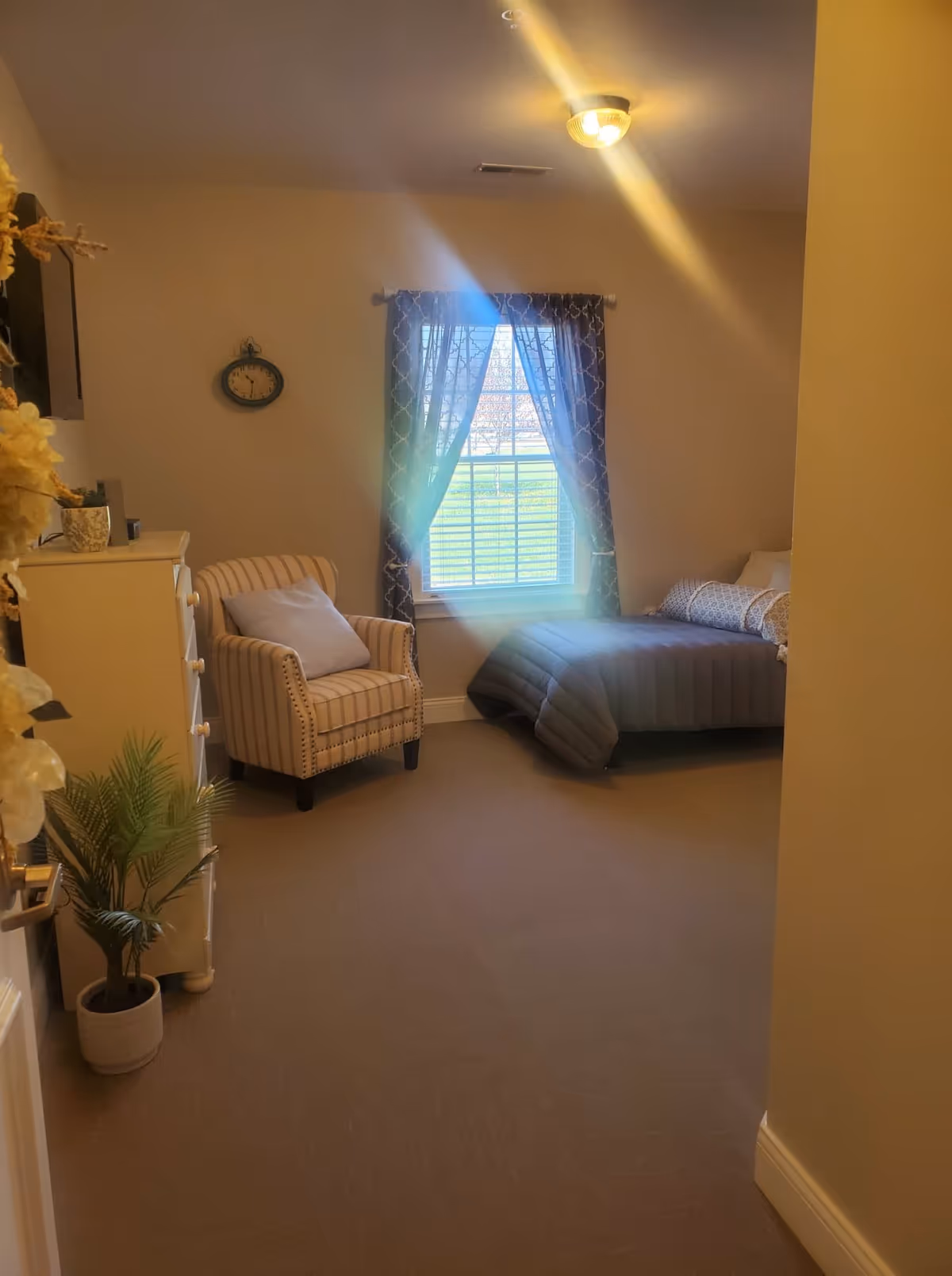A cozy bedroom with a single bed covered in a gray comforter and patterned pillows, a striped armchair with a cushion, a white dresser with decorative items, a potted plant on the floor, and a window with patterned curtains letting in natural light.