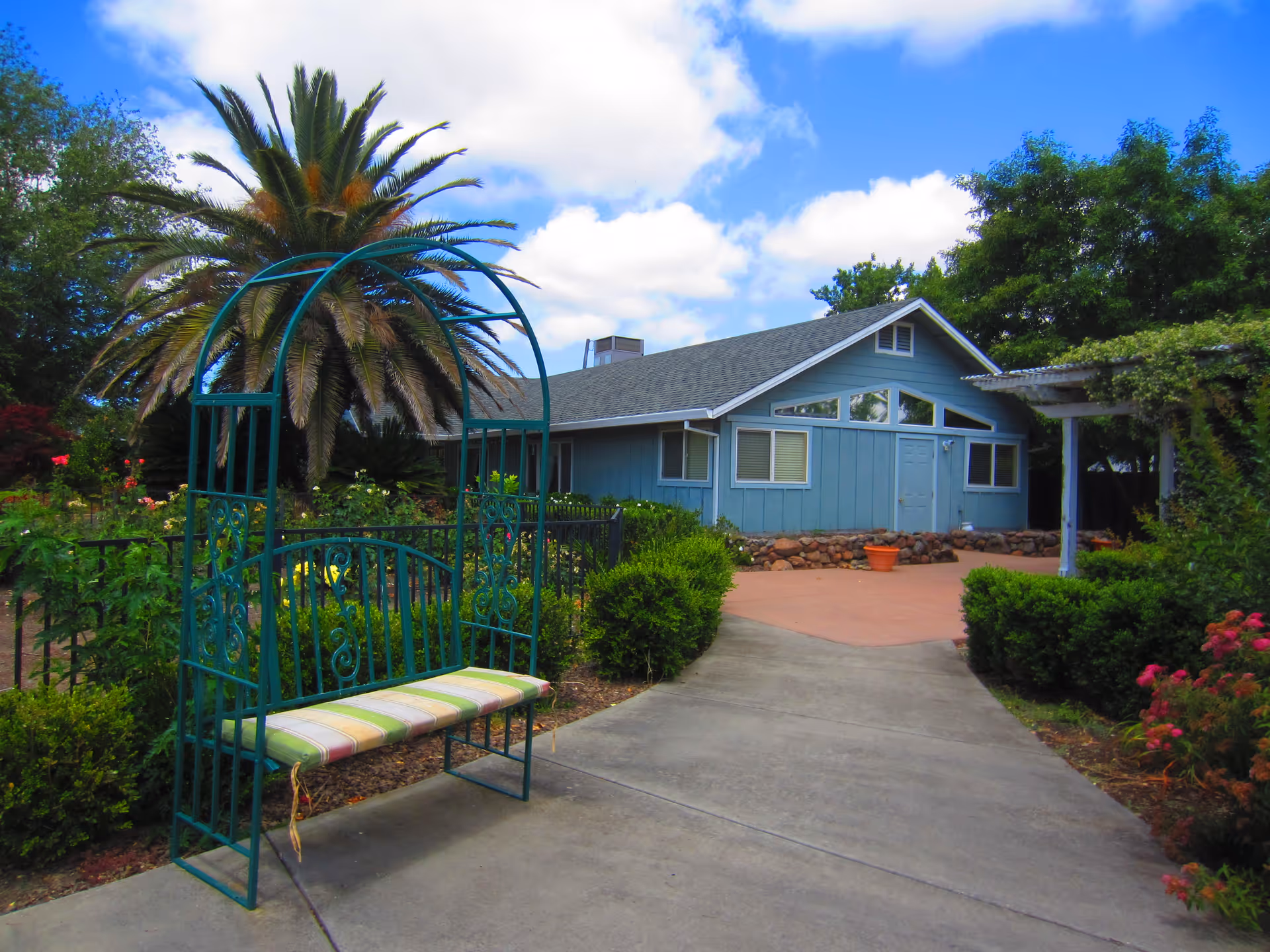 Outdoor garden area with a green metal bench that has a striped cushion, a large palm tree, various bushes and flowers, and a blue building with white trim in the background under a partly cloudy sky.