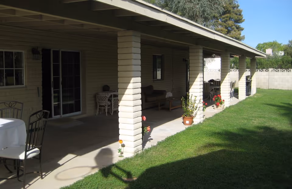 Covered patio area with brick columns, outdoor furniture including tables and chairs, potted plants, and a grassy yard extending alongside the patio under a clear blue sky.