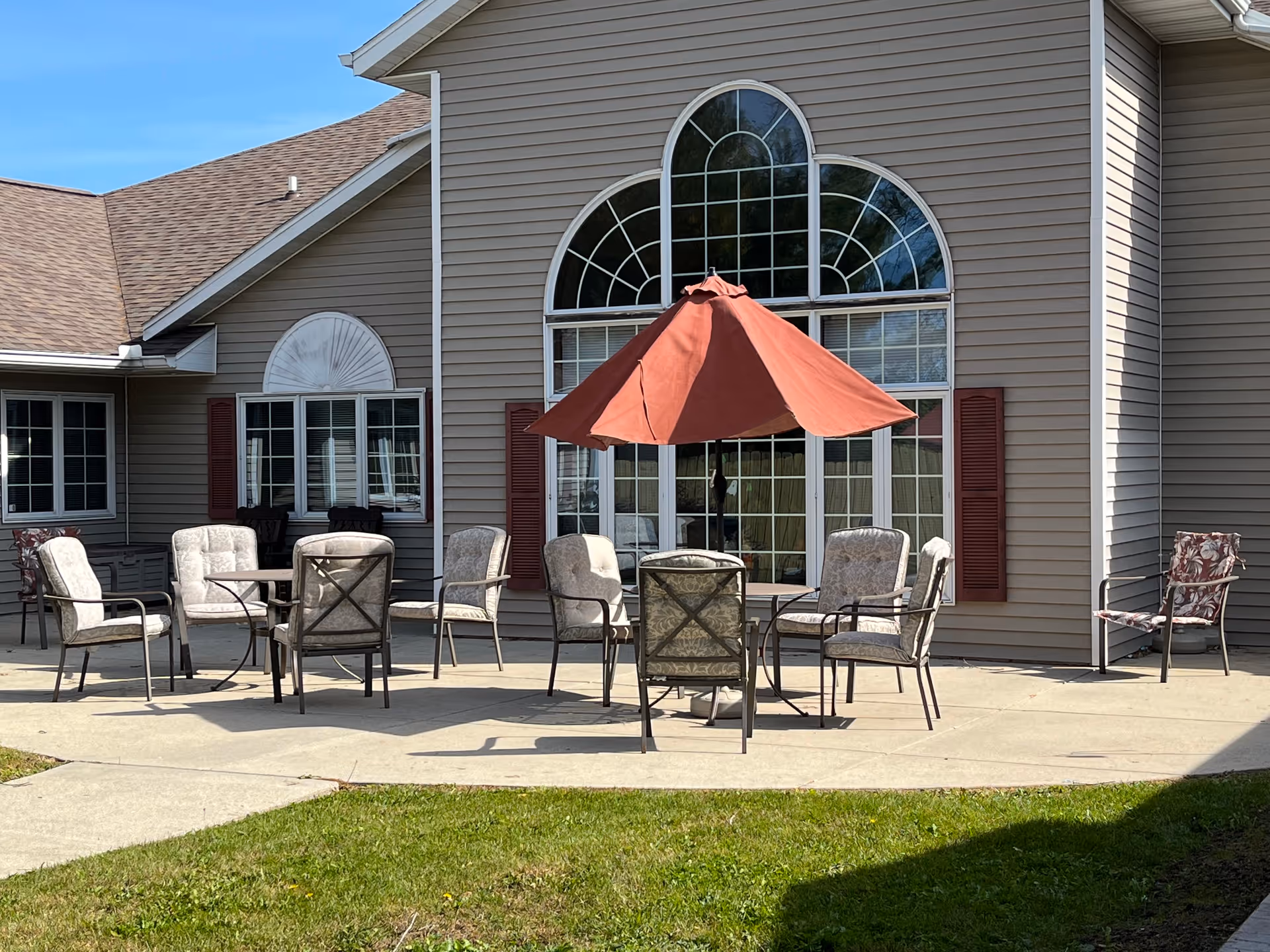 Outdoor patio area with multiple cushioned chairs arranged around two tables, one of which has a large red umbrella. The patio is adjacent to a building with beige siding and large decorative windows with red shutters.